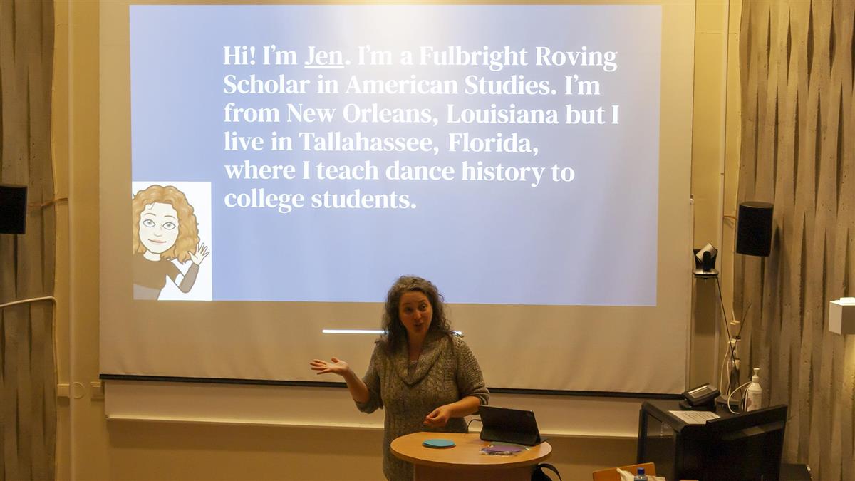 Jen Atkins fra Fulbright holder foredrag om amerikansk samfunn og kultur på auditoriet til studiested Solhaugen. Hun veiver med armene og er engasjert mens hun introduserer seg selv.  På lysbildet bak henne står det: "Hi! I'm Jen. I'm a Fulbright Roving Scholar in American Studies. I'm from New Orleans, Louisiana but I live in Tallahassee, Florida, where I teach dance history to college students." - Klikk for stort bilde