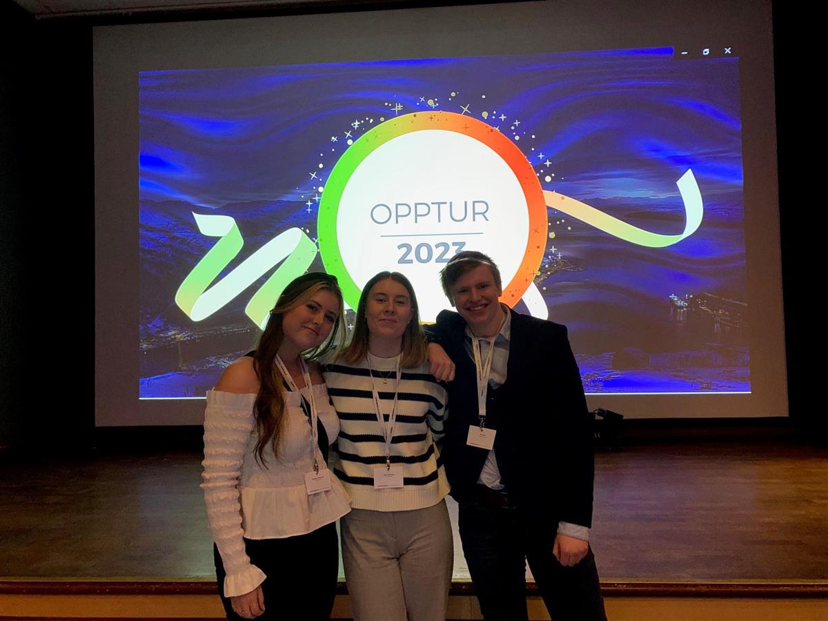 Tre elever poserer foran scenen med Opptur 2023-logoen synlig på skjerm bak dem. - Klikk for stort bilde